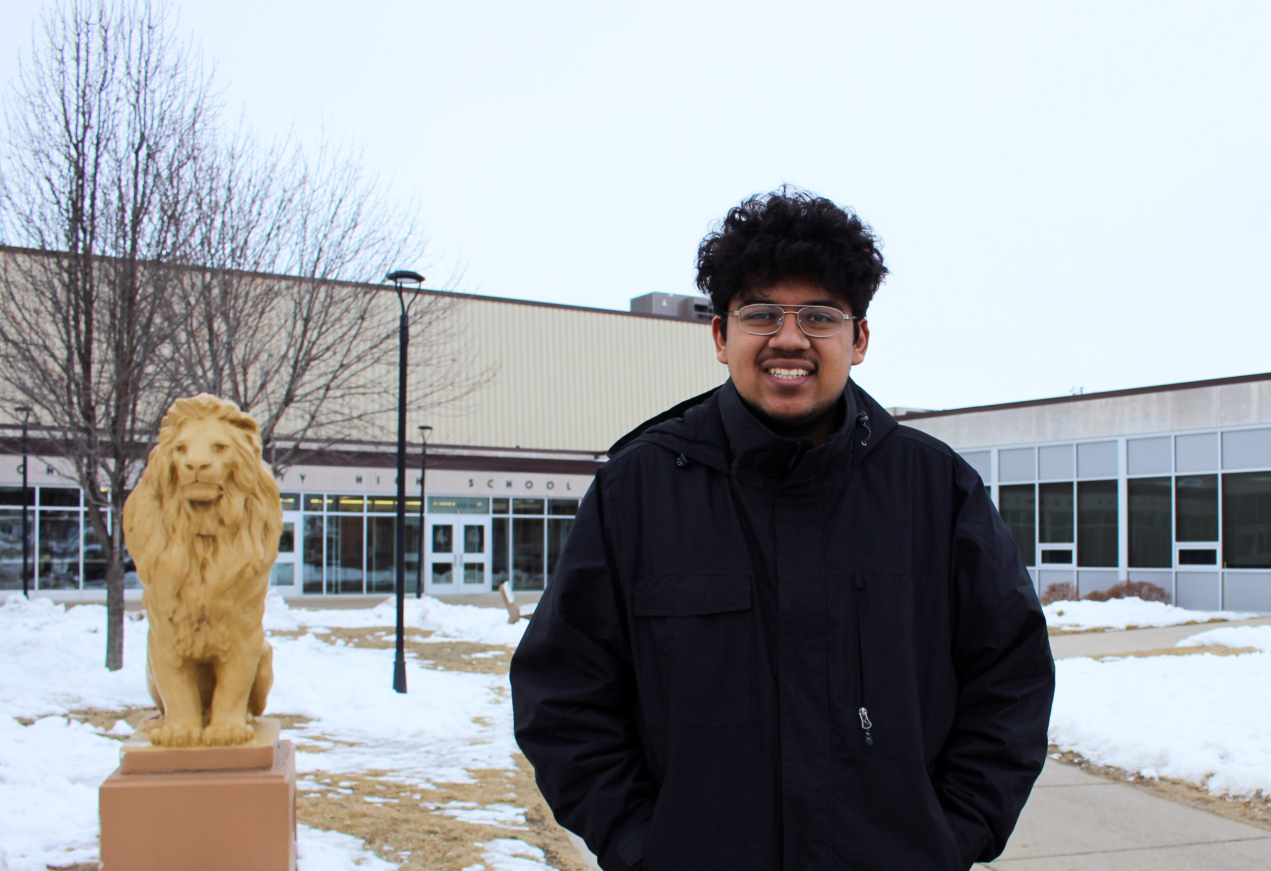 A young man in a black jacket with his hands in his pants pocket to the right of a sculpture of a lion with shops in the background after a heavy snowfall.