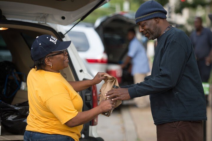 A woman next to the open trunk of a car hands a paper bag to a man who receives it with both hands.