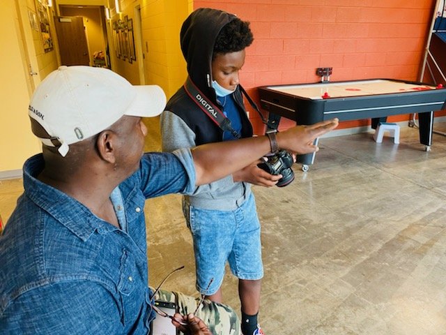 A man with a cell phone pointing in the front direction next to a child wearing a face mask and holding a camera.